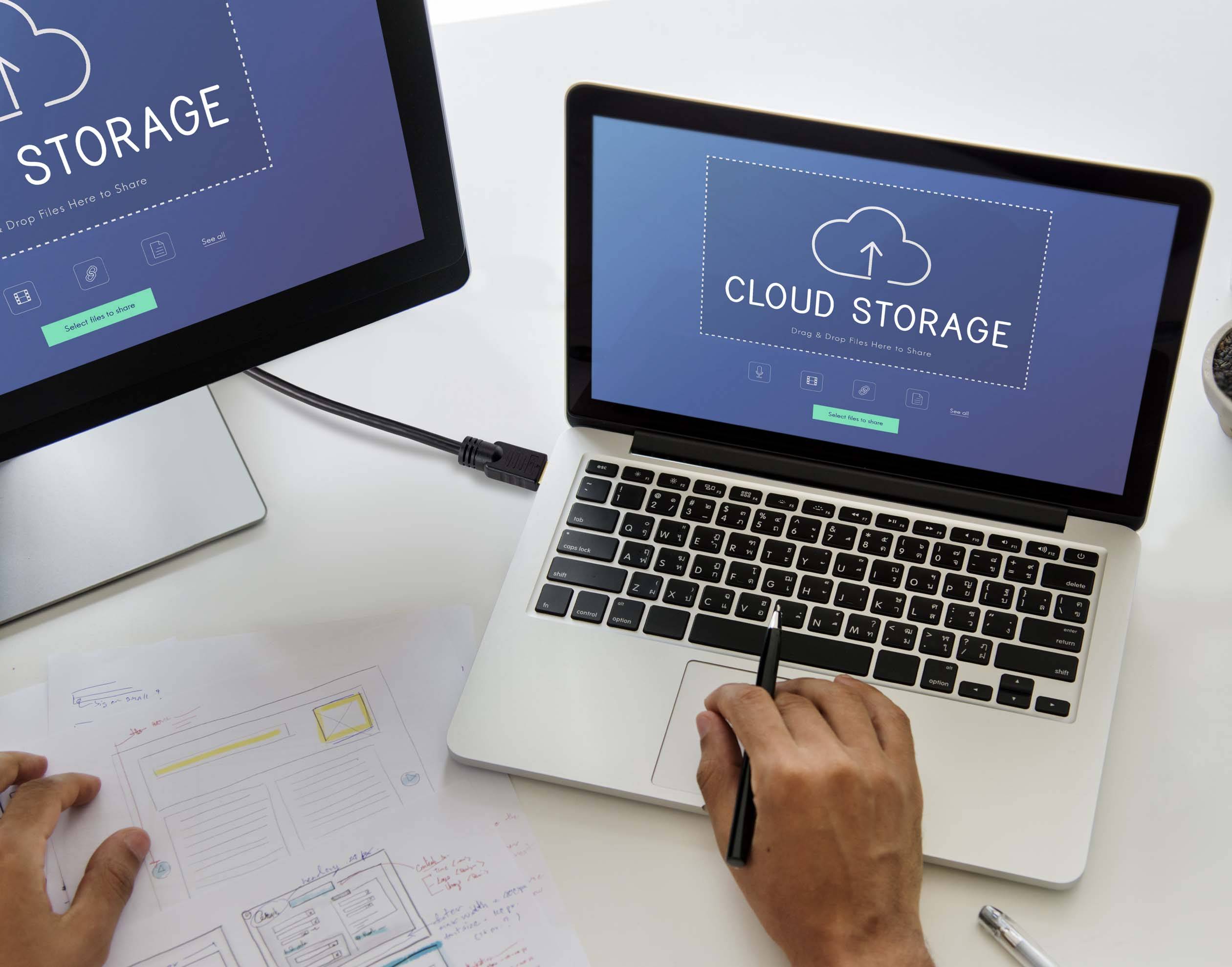 Na dvou obrazovkách je zobrazeno 'Cloud Storage', vedle sedící osoba si současně dělá poznámky na papíry. Téma: digitální ukládání a správa dat.