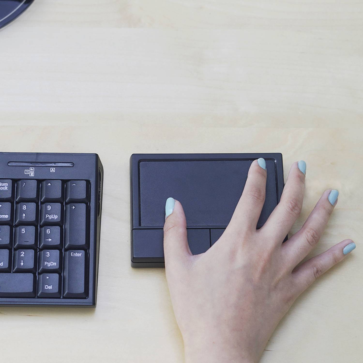 Ruka ovládá grafický tablet vedle kompaktní klávesnice na dřevěném stole, což naznačuje kreativní digitální práci.