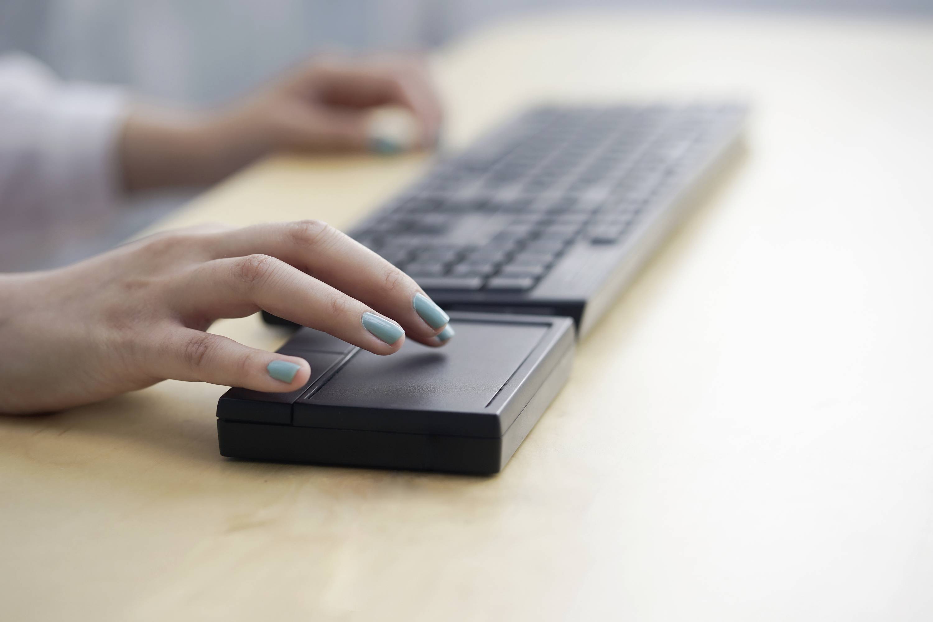 Ruka s modrými nehty ovládá touchpad černého laptopku na pracovním stole, v pozadí klávesnice.