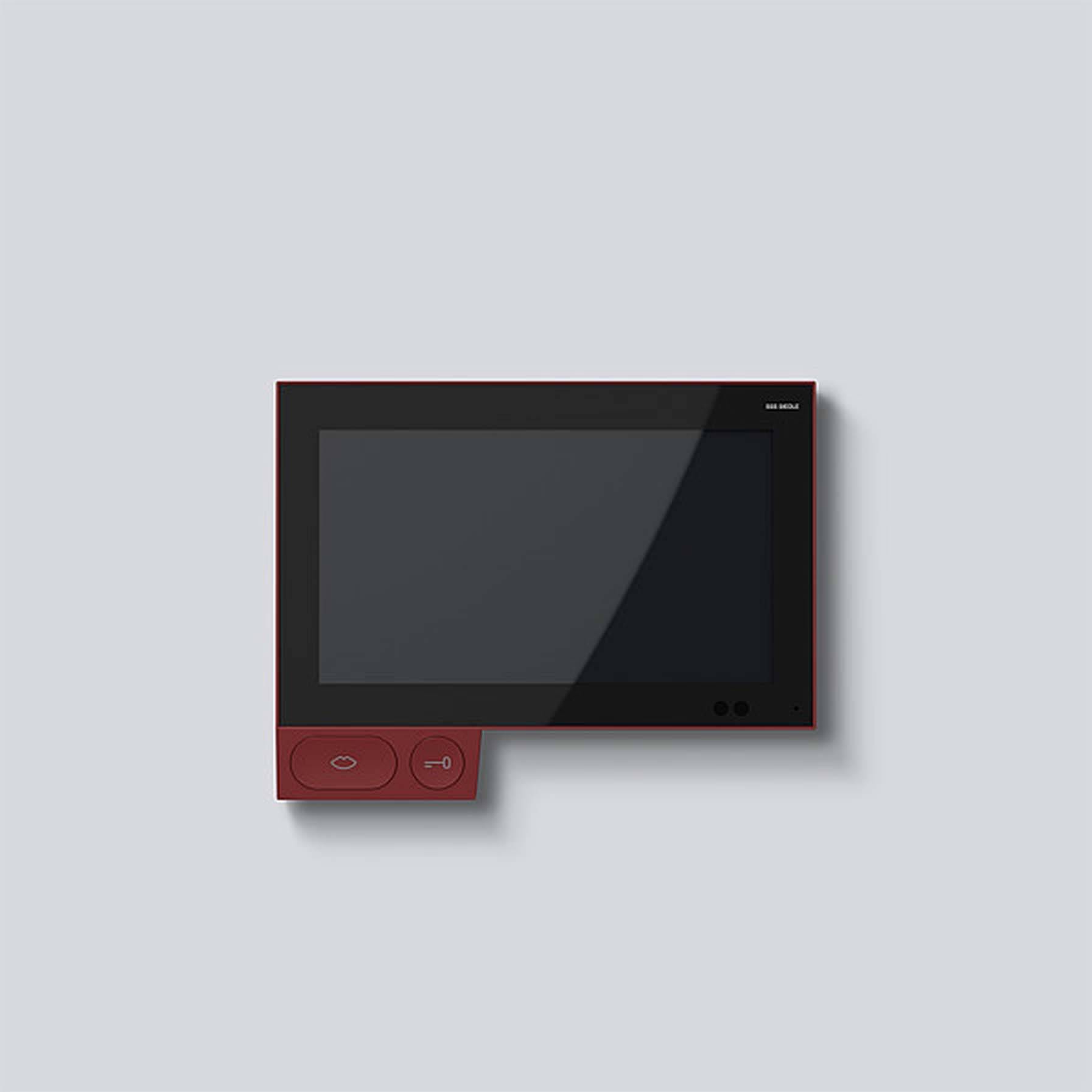 Červené elektronické zařízení s displejem a dvěma tlačítky, připevněné na stěně. Má minimalistický design.