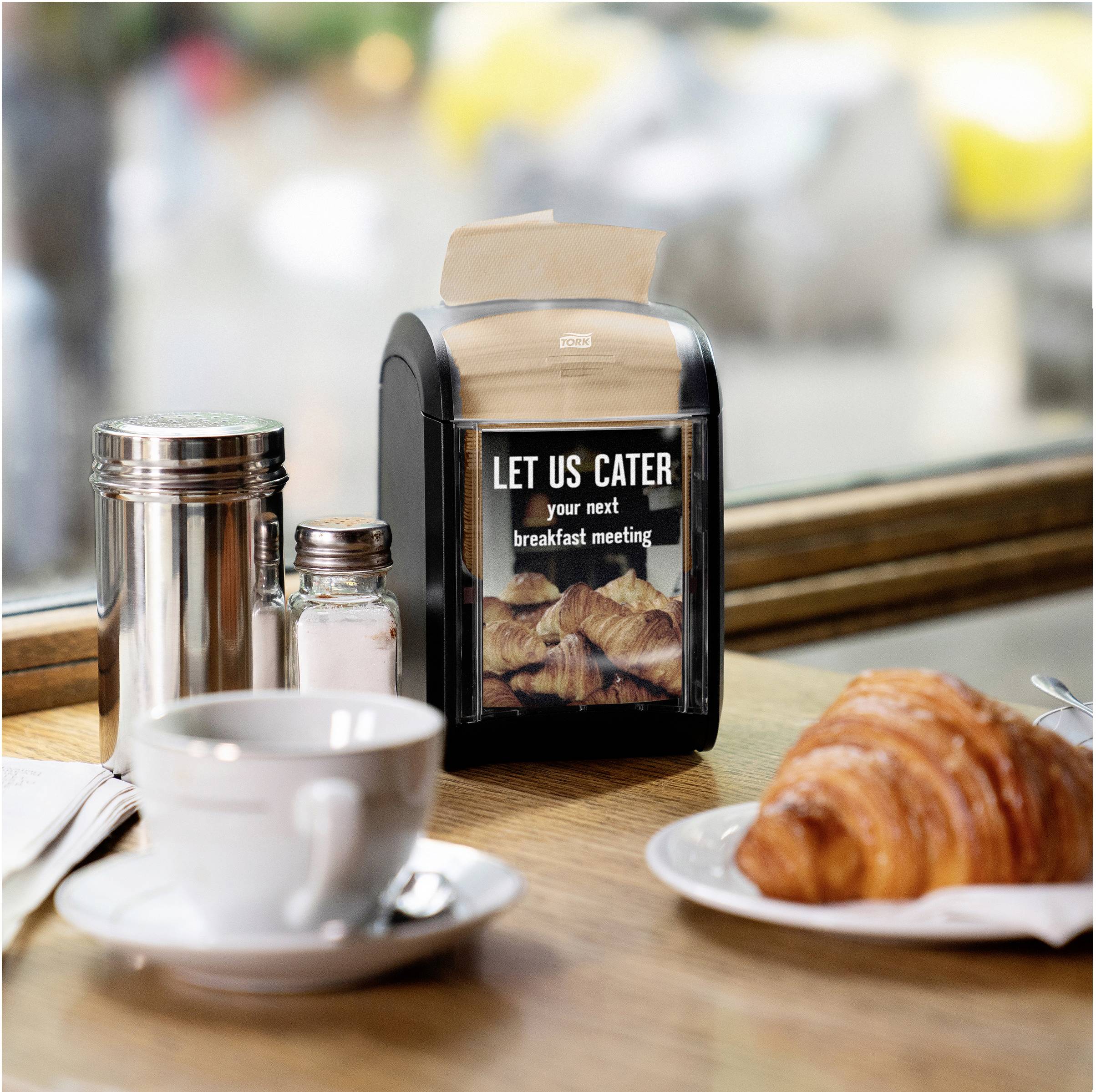 Stůl v kavárně s croissantem, šálkem kávy, slánkou a cukřenkou. Uprostřed stojí displej s nápisem 'LET US CATER your next breakfast meeting'.