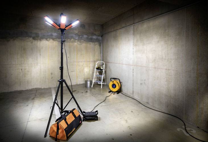 Jasný stavební reflektor osvětluje prázdný betonový prostor. Žebřík a navinutý prodlužovací kabel jsou opřeny o zeď.