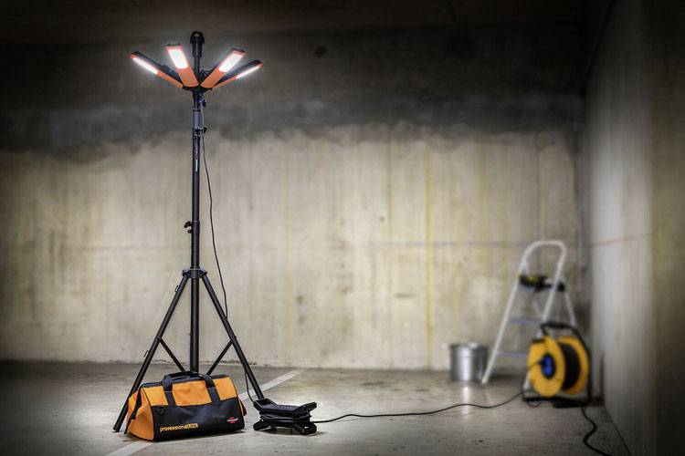 Na obrázku je vidět stavební reflektor na stativu v suterénu. Vedle něj leží taška s nářadím a prodlužovací kabel.