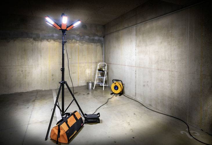 Osvětlený prostor s bouracími reflektory, oranžovou látkovou brašnou na nářadí, žebříkem a kabelovým bubnem na betonovém podkladu.