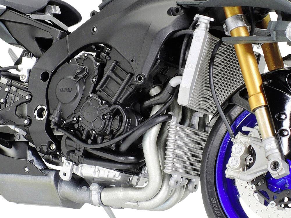 Detailní záběr na oblast motoru a předního kola motocyklu, zobrazující motorový blok, zlatý tlumič a modré kolo.