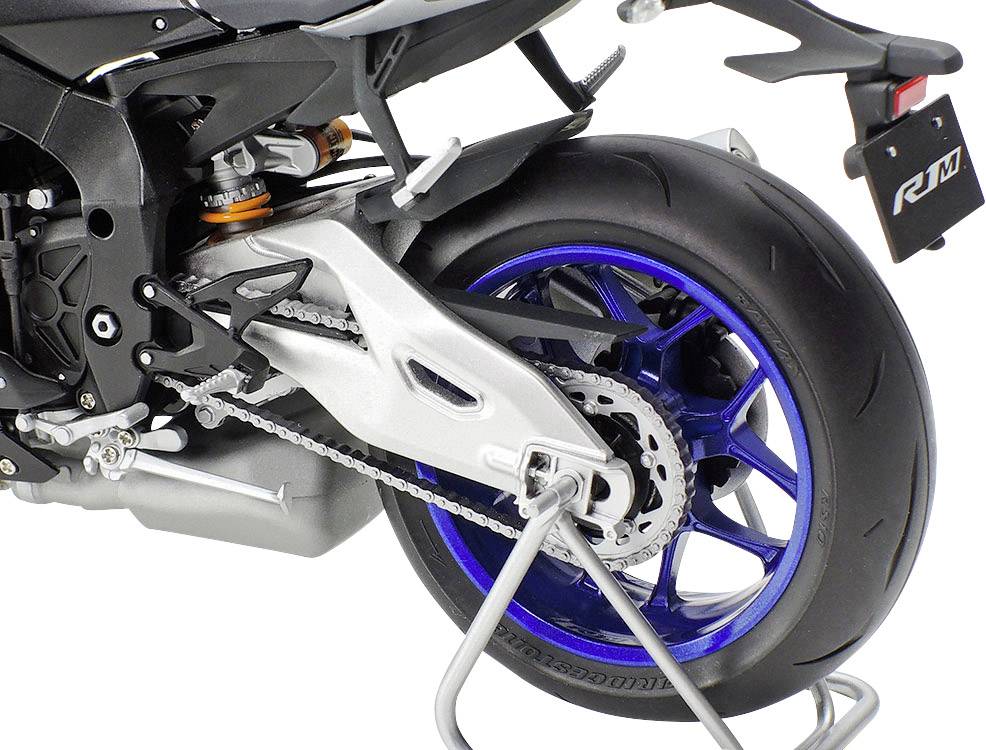 Detailní záběr zadního kola motocyklu s modrými ráfky, řetězem a výfukem; stojí na stojanu.
