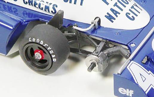Detailní záběr modrého závodního pneumatiky s nápisem 'Goodyear'. Pneumatika je namontována a zobrazuje části zavěšení kola.