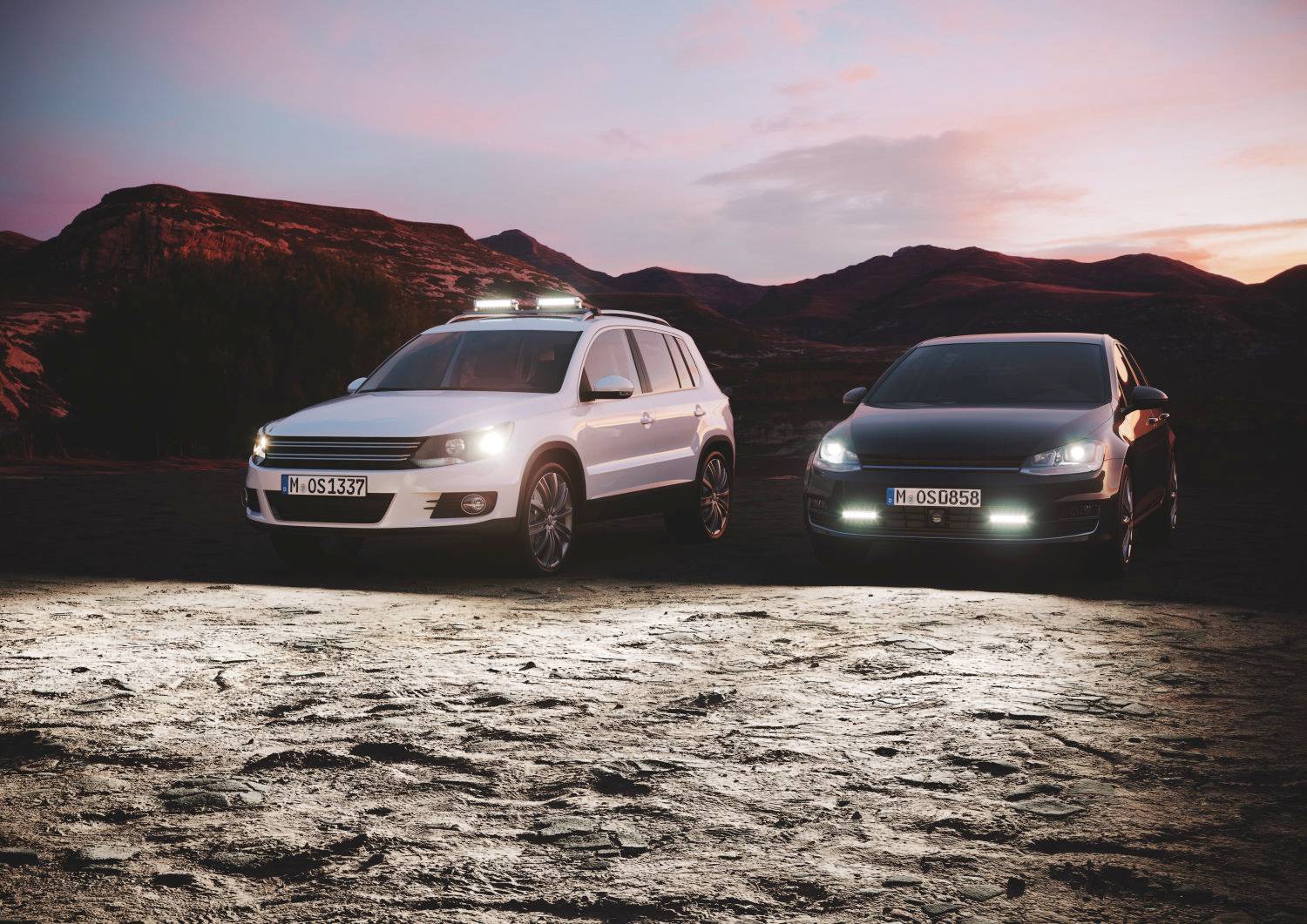 Dva automobily stojí vedle sebe v pouštní krajině za soumraku; vlevo bílý, vpravo černý se světly reflektorů.