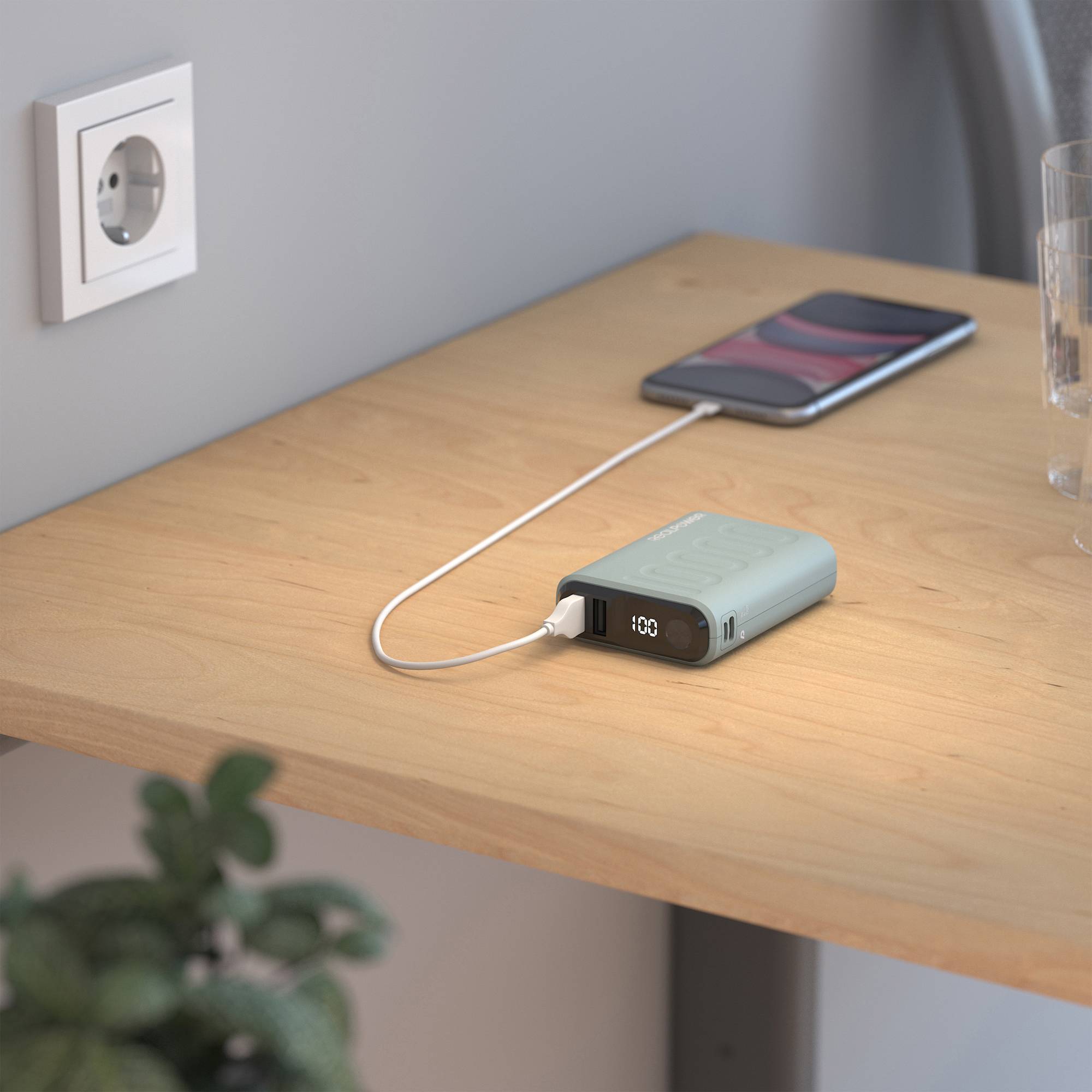 Na dřevěném stole se nabíjí smartphone pomocí powerbabanky. V pozadí je viditelná elektrická zásuvka a sklenice vody.