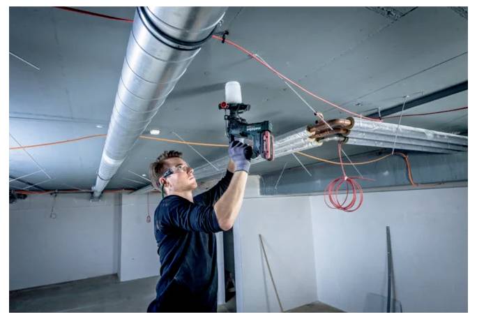 Osoba v bezpečnostních brýlích používá elektrické nářadí k instalaci potrubí v stropu, obklopena elektrickými vodiči v nedokončené místnosti.