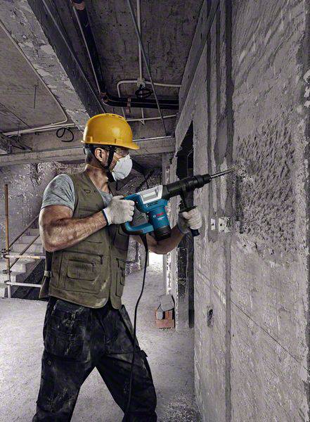 Dělník v ochranném oděvu a helmě vrtá do betonové stěny na staveništi. Při vrtání vzniká prach. V pozadí jsou schody a betonové stěny.