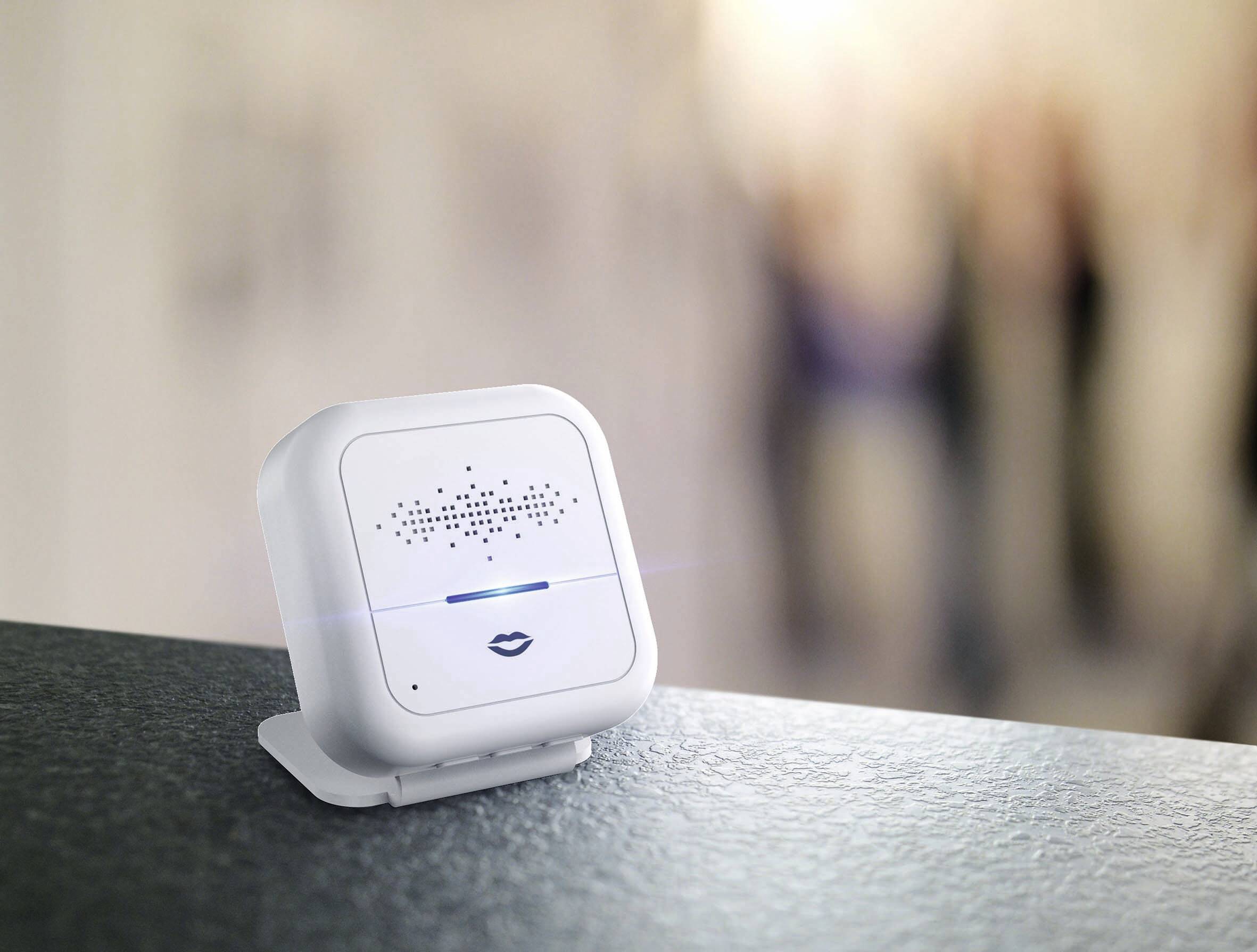 Kompaktní bílé zařízení pro hlasové ovládání stojí na stole. V pozadí jsou rozmazané osoby.