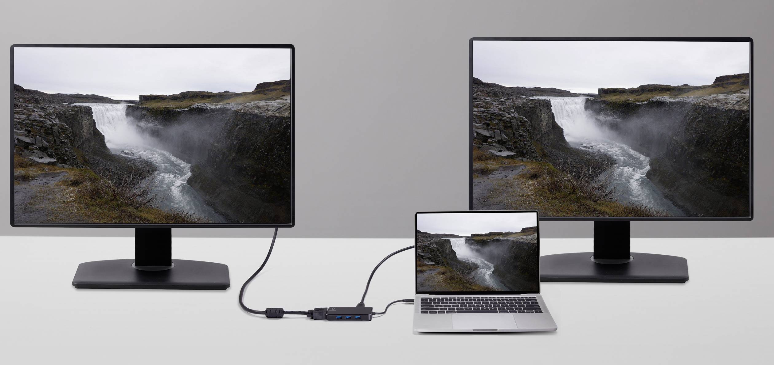 Notebook je připojen přes adaptér ke dvěma monitorům, které zobrazují stejný krajinný obraz vodopádu.