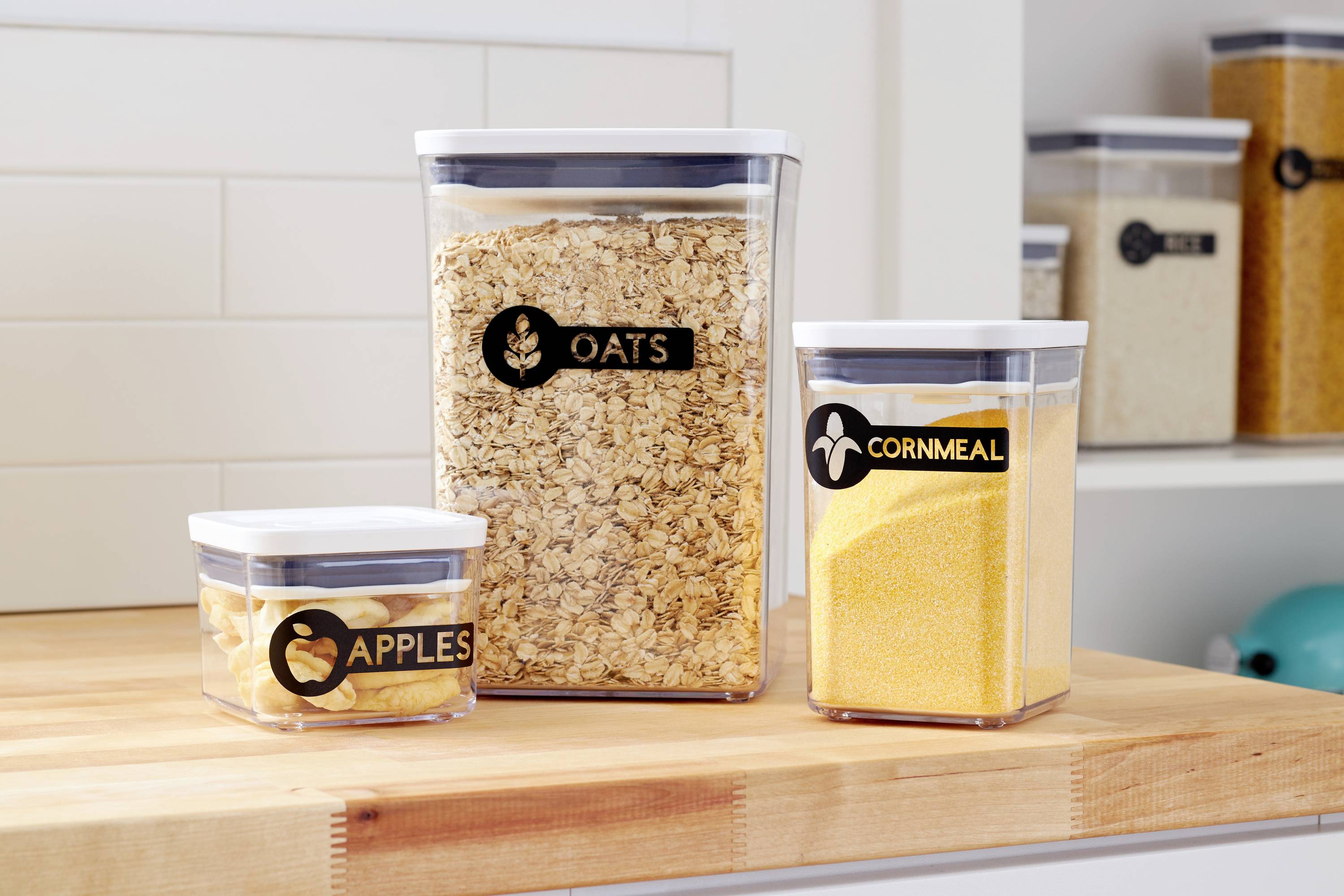 Tři průhledné skladovací nádoby na dřevěném povrchu, naplněné jablky, ovesnými vločkami a kukuřičnou moukou, každá opatřená štítkem.