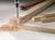 Vrtačka vrtá díru do dřevěného trámu na pracovním stole, obklopená dřevěnými pilinami. Několik kusů dřeva leží vedle.