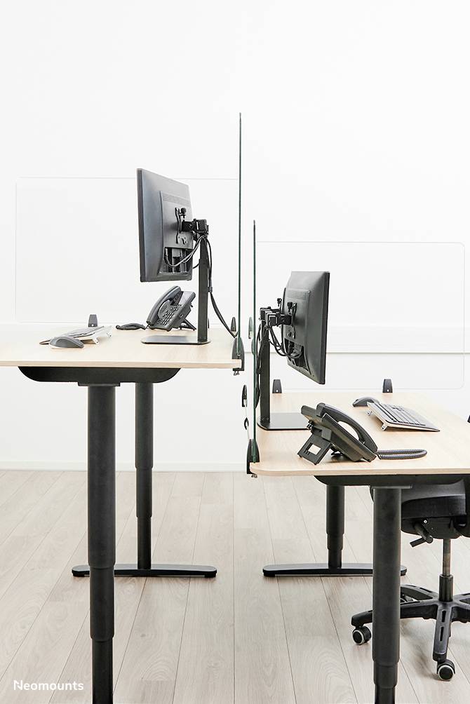 Dva výškově nastavitelné psací stoly s monitory a telefony, oddělené průhlednou přepážkou, na dřevěné podlaze.