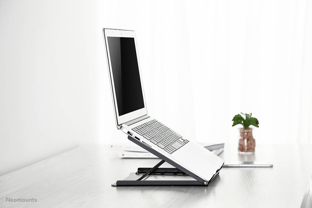 Laptop stojí na nastavitelném stojanu na psacím stole. V pozadí je viditelná rostlina.