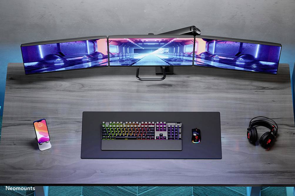 Pracovní stůl s trojitým nastavením monitorů, podsvícenou klávesnicí, myší, smartphonem a sluchátky; ideální pro hraní her.