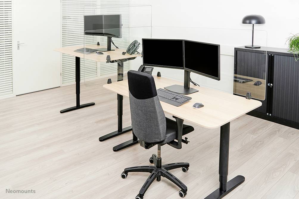 Dva psací stoly s monitory, klávesnicemi a kancelářskými židlemi v moderní kanceláři. V pozadí skříň a lampa.