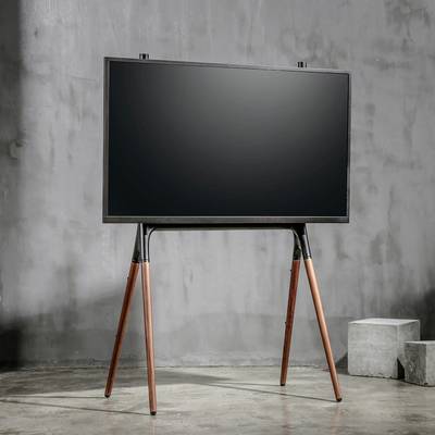 Moderní televize stojí na trojnožkovém dřevěném stojanu v minimalistickém prostoru s betonovými stěnami.