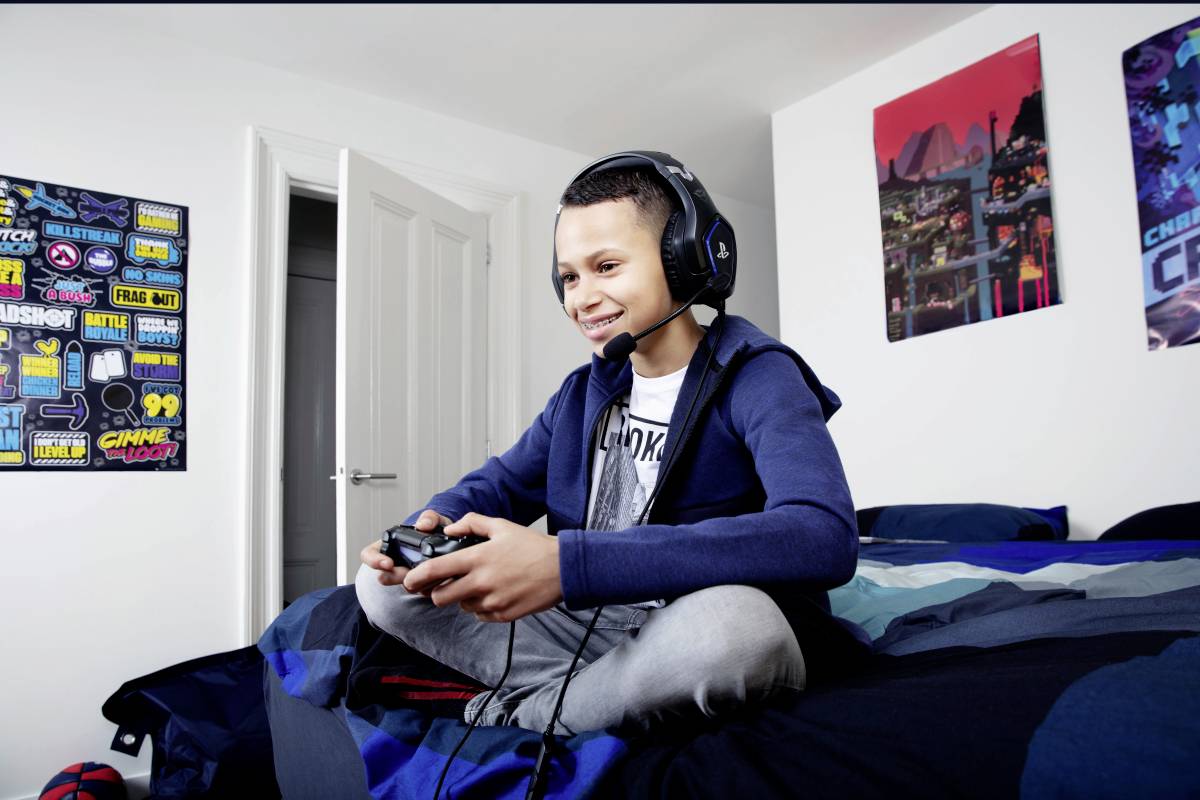 Chlapec hraje videohru ve své ložnici, má nasazená sluchátka, sedí na posteli, v pozadí světlé stěny zdobené plakáty.