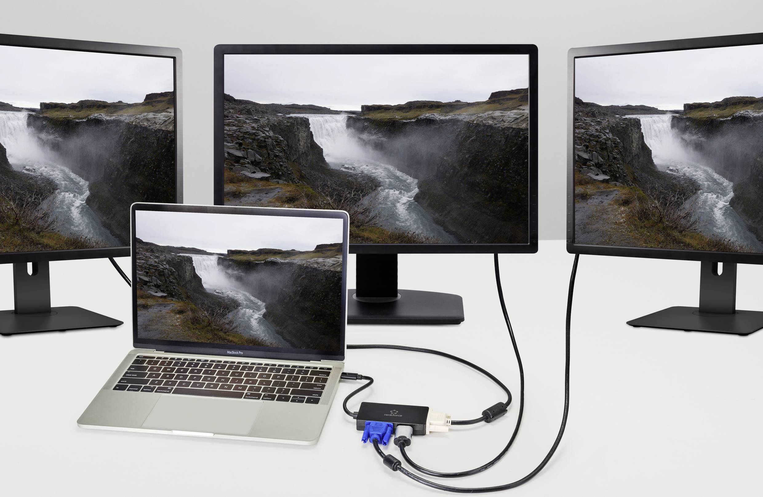 Notebook je připojen ke dvěma monitorům, které zobrazují stejné pozadí s krajinou s řekou, propojené adaptéry a kabely.