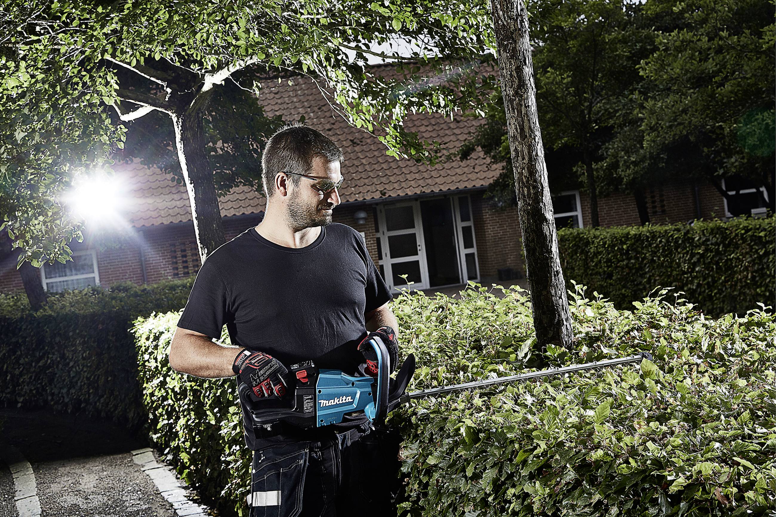 Muž stříhá živý plot elektrickým plotostřihem na zahradě. Je slunečno a má na sobě ochranné brýle a rukavice.