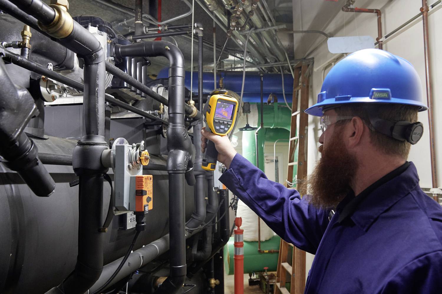 Technik v ochranných brýlích a helmě kontroluje teplotu potrubí pomocí infračerveného teploměru v průmyslovém prostoru.