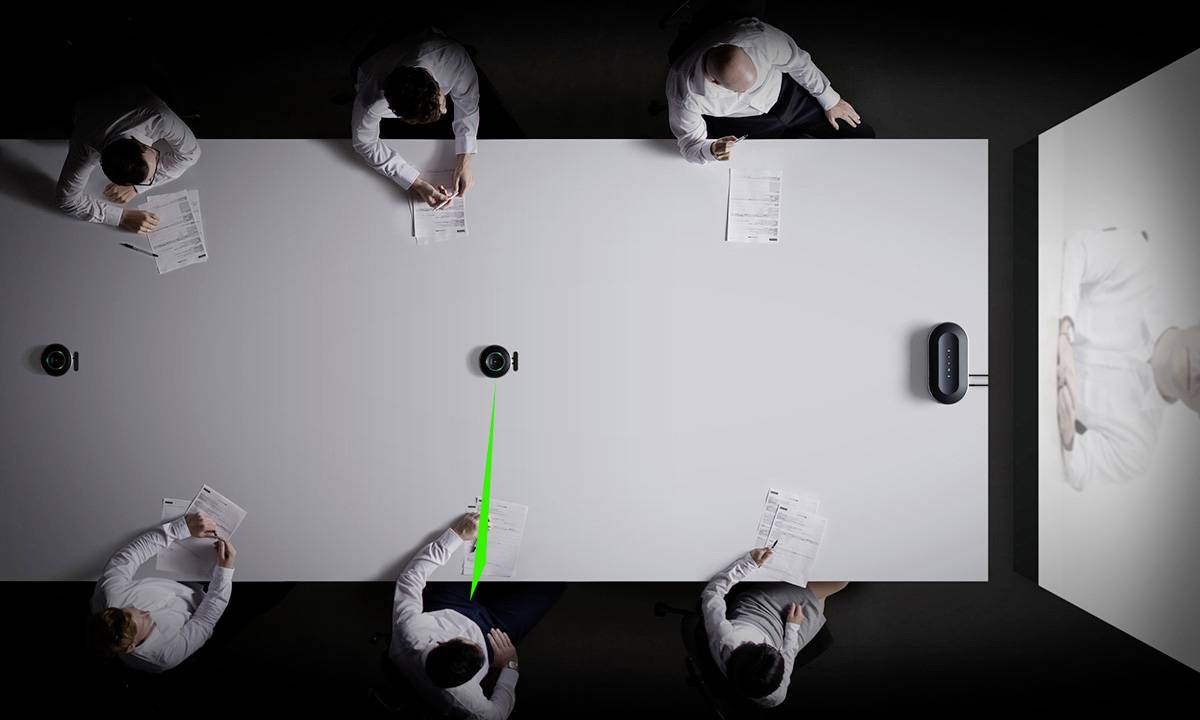 Šest osob v bílých košilích sedí u obdélníkového konferenčního stolu a sleduje videoprezentaci. Zelená čára něco vyznačuje na stole.