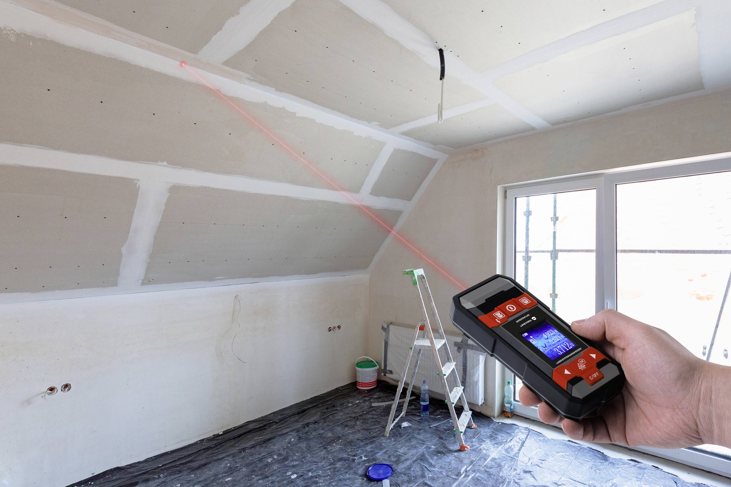 Osoba měří výšku místnosti renovované místnosti laserovým dálkoměrem. V pozadí stojí žebřík.