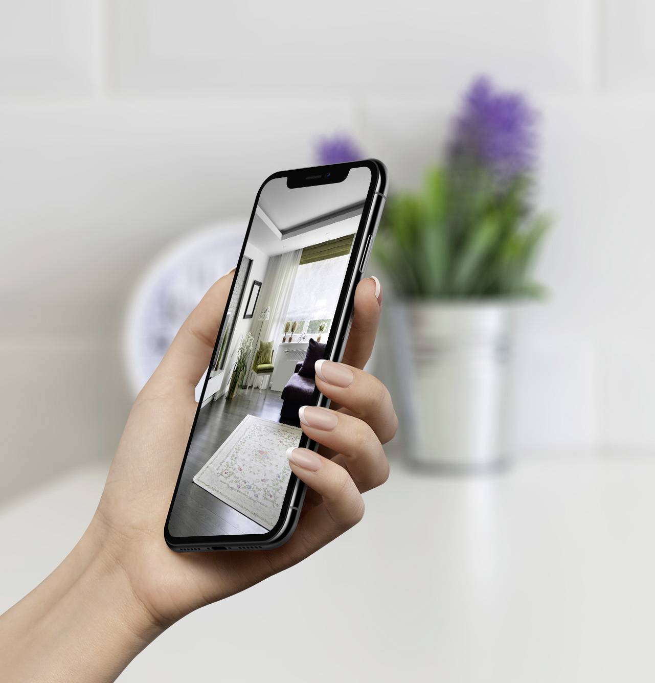 Jedna ruka drží smartphone s fotografií moderně zařízeného obývacího pokoje. V pozadí je rozmazaně viditelný květináč.