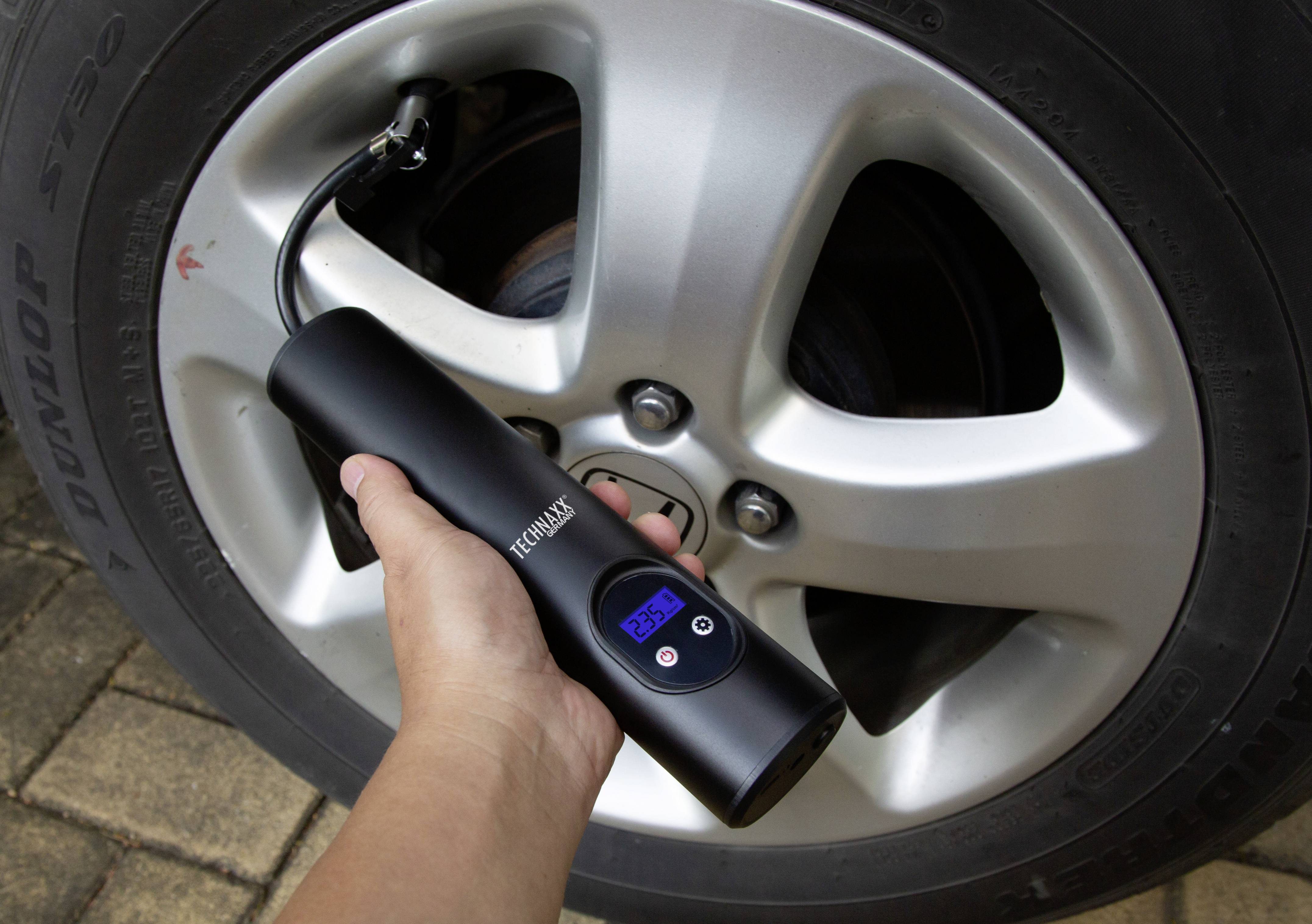 Ruční zařízení měří tlak pneumatiky automobilu. Displej zobrazuje '2.55'. Pneumatika stojí na dlážděném povrchu.