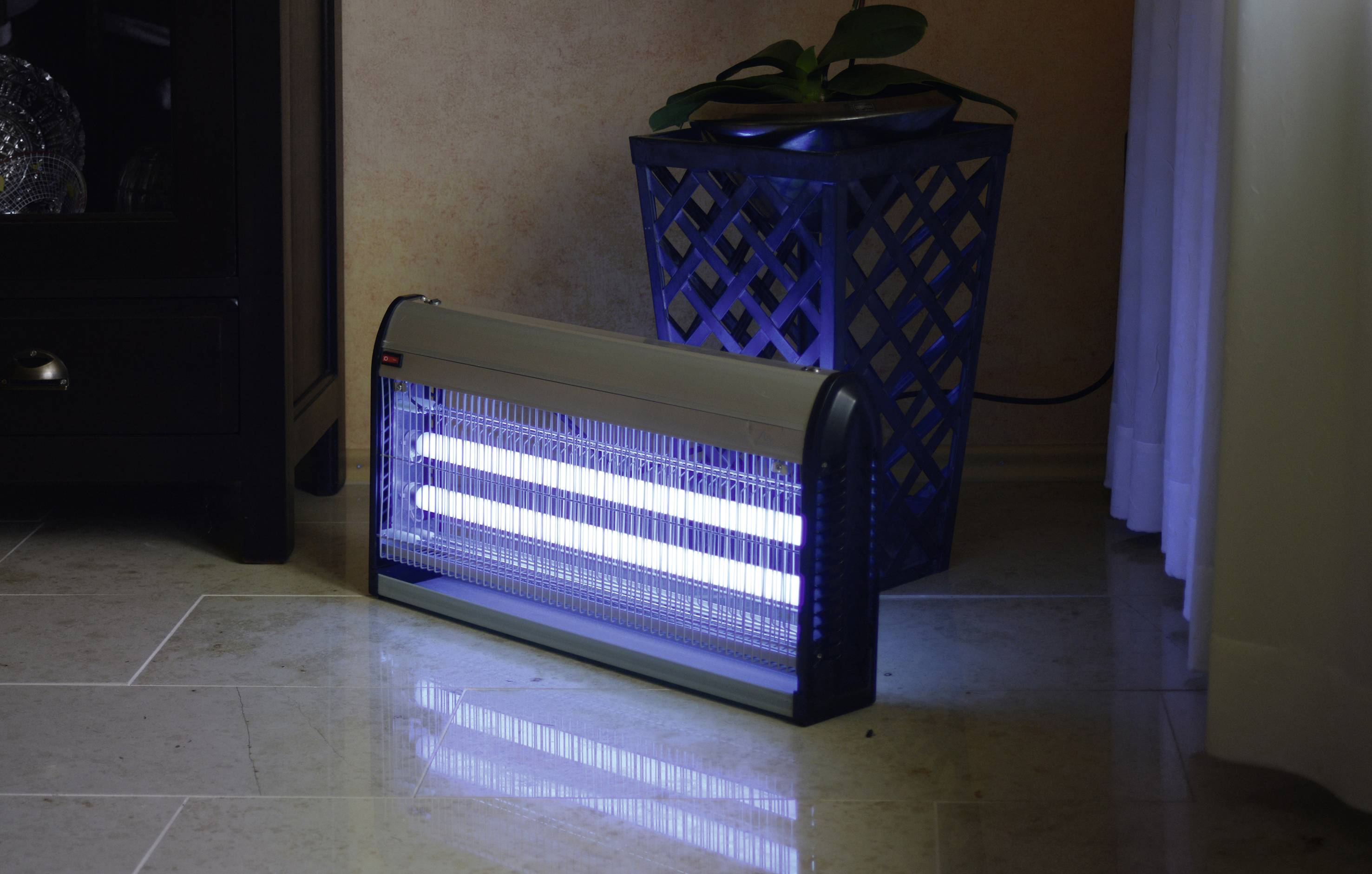 Elektrický lapač hmyzu s modrým světlem stojí na dlaždicové podlaze vedle květináče.