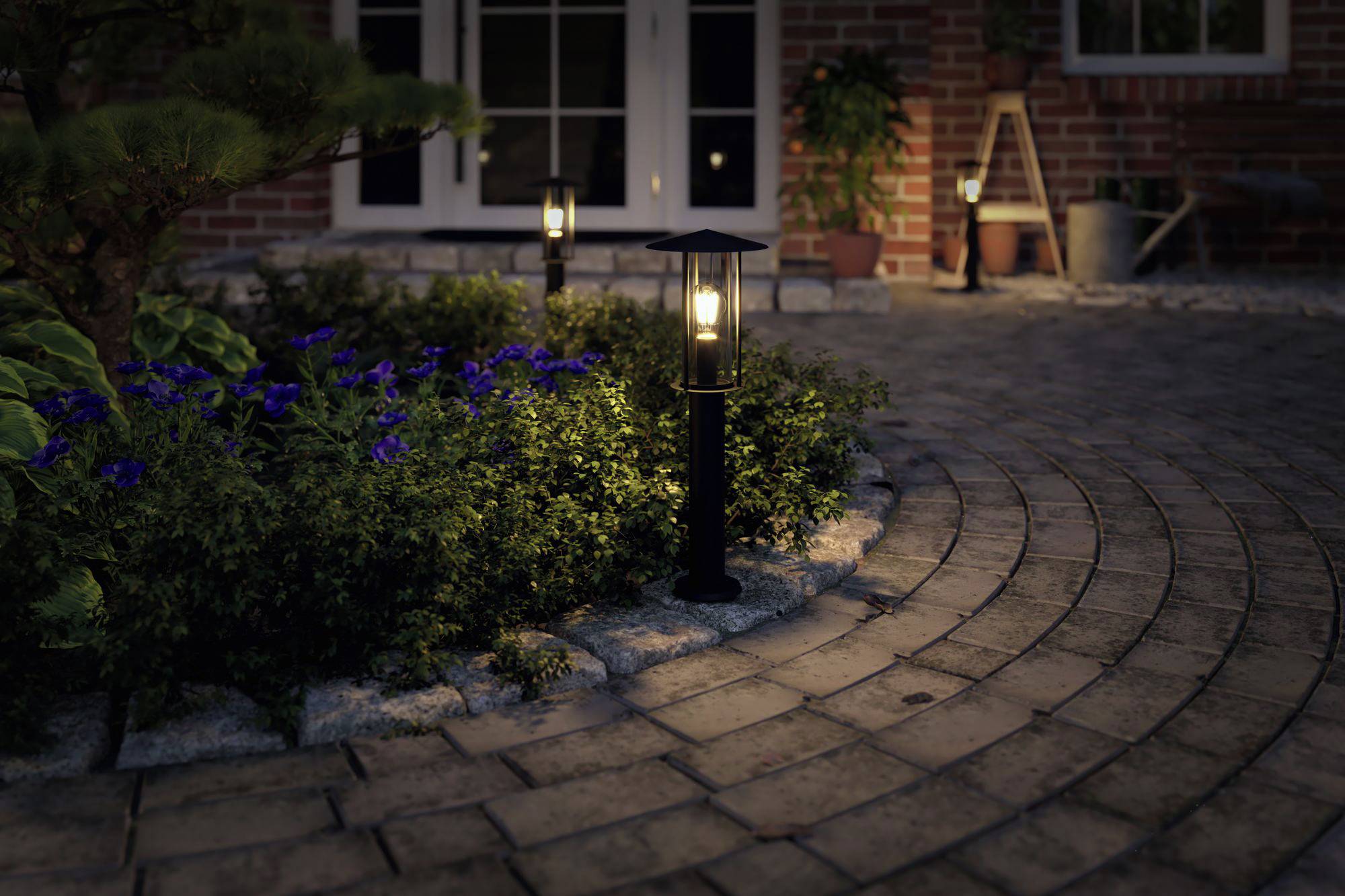 Zahradní cesta s dlažbou z mozaikových kostek, lemovaná rozkvetlými rostlinami a třemi zářícími lampami. Dveře a rostliny v pozadí.
