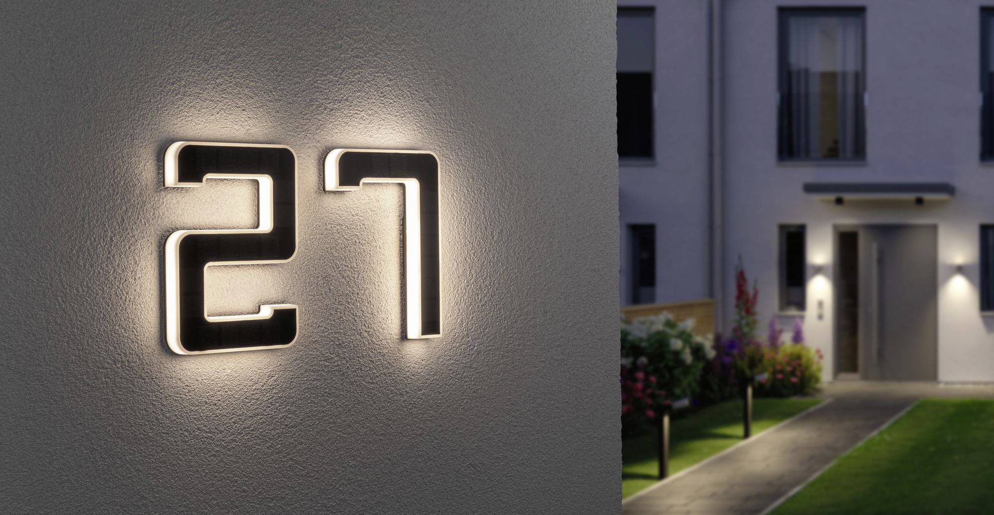 Osvětlené číslo domu '27' na zdi domu. V pozadí vede osvětlená cesta k moderní budově.