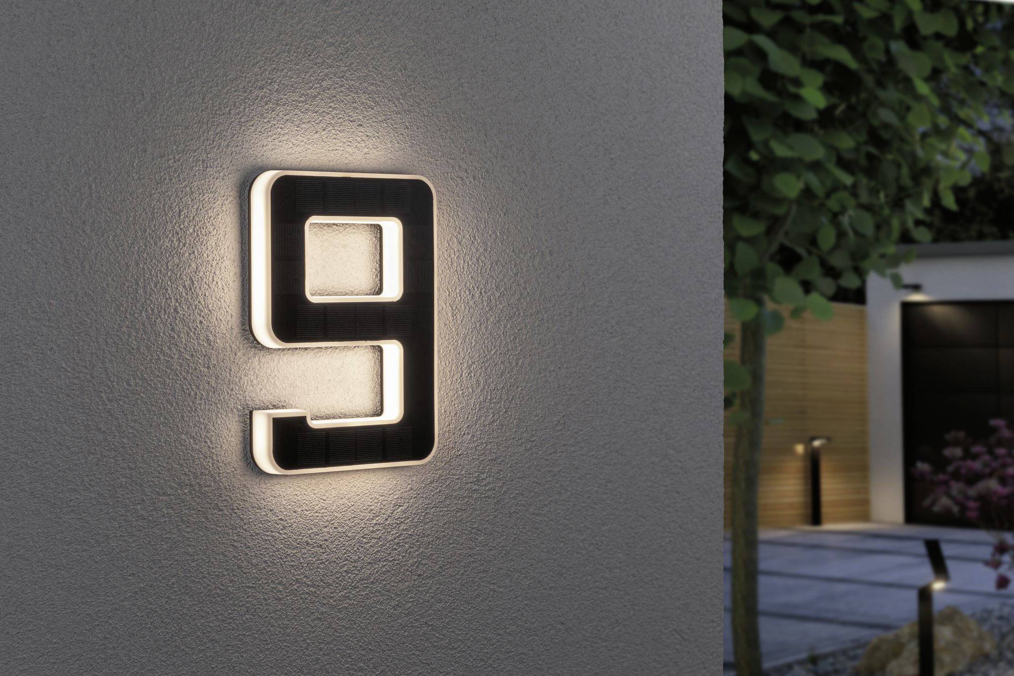 Osvětlené číslo domu '9' na zdi večer. V pozadí rozmazané stromy a vstup do domu.