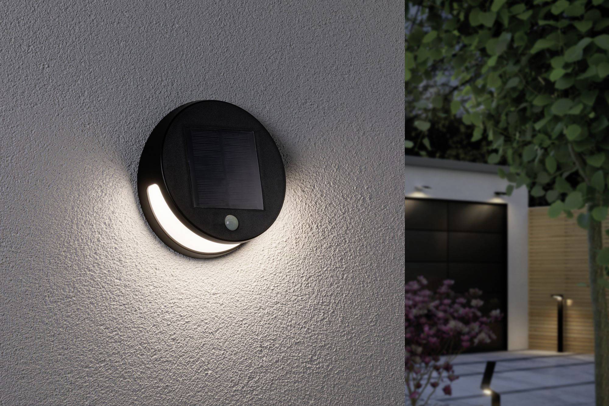 Kulatá nástěnná lampa se solárním panelem a pohybovým senzorem osvětluje v noci zahradní prostor. V pozadí jsou viditelné stromy.