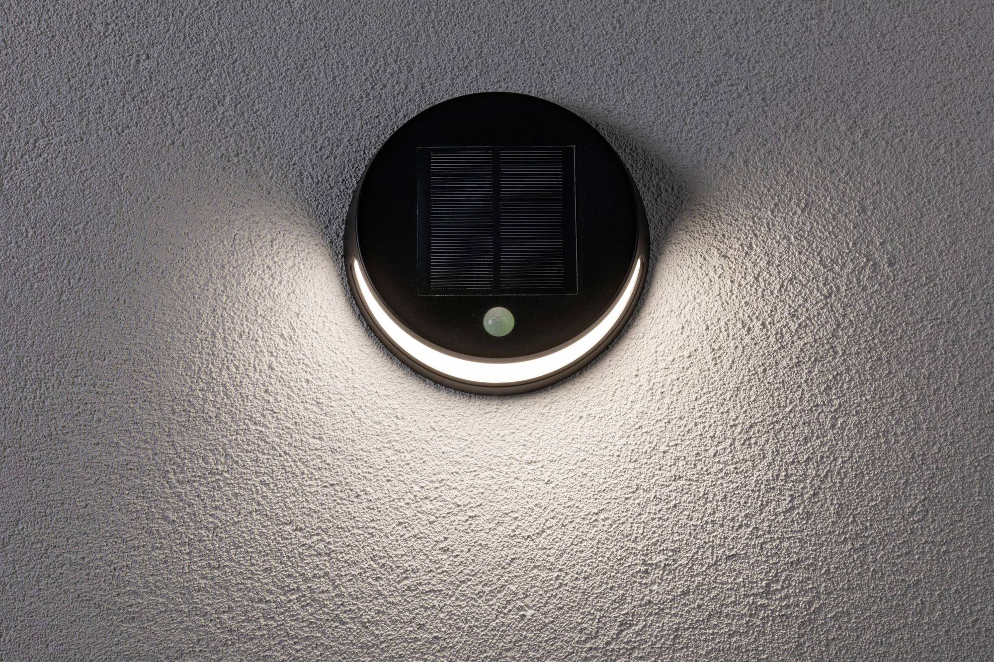 Kulatá solární lampa na šedé zdi svítí a její světlo se šíří nahoru a dolů.
