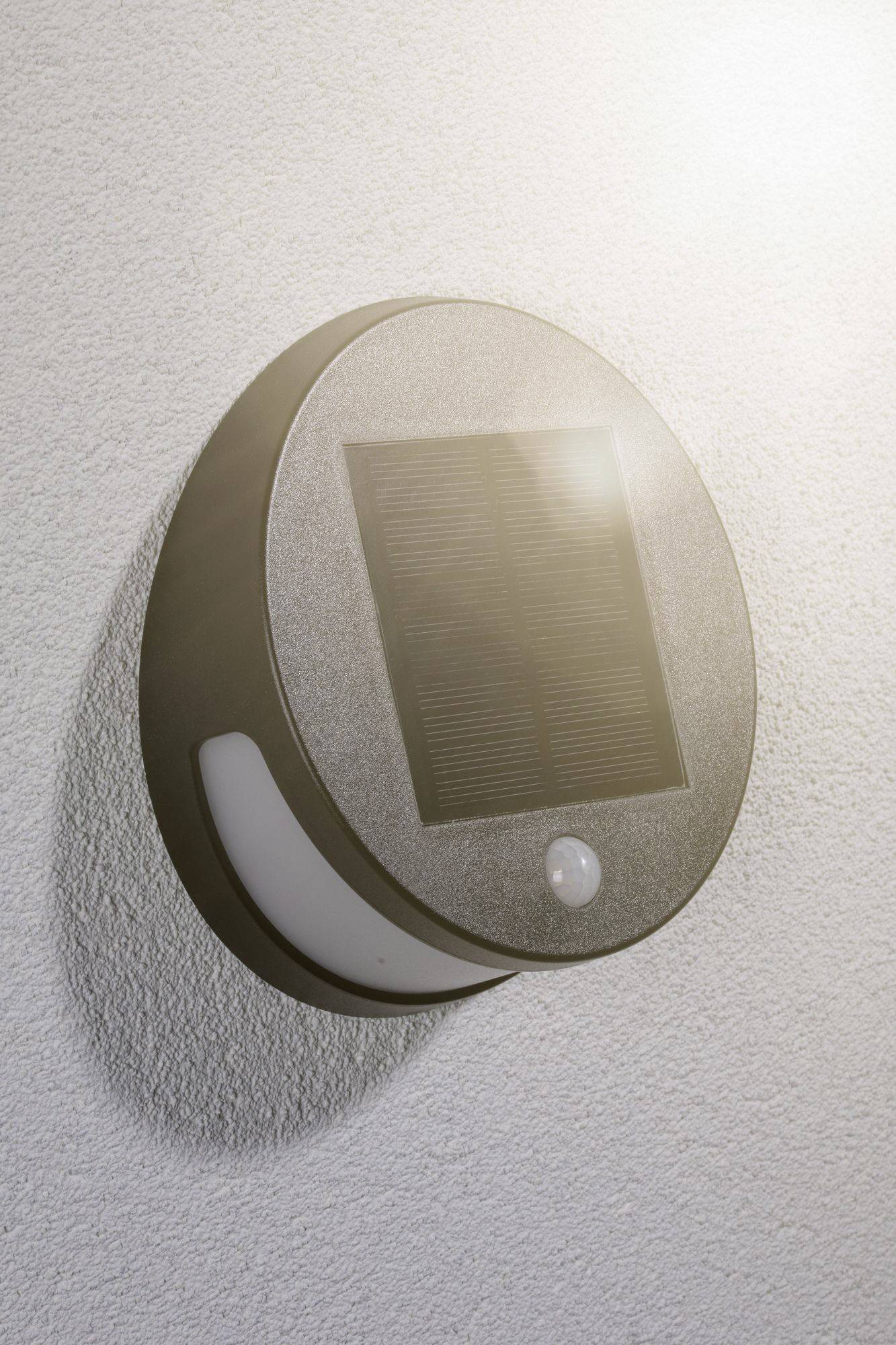 Kulatá solární lampa s integrovaným solárním článkem a senzorem na zdi, osvětlená slunečním světlem.