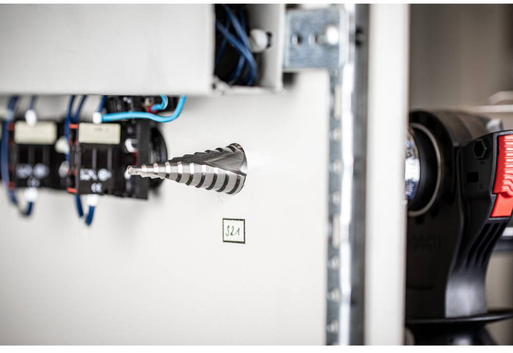 Průmyslová rozvodná skříň s namontovaným stupňovitým vrtákem a elektrickým vedením. Vpravo jsou patrné části nástroje.