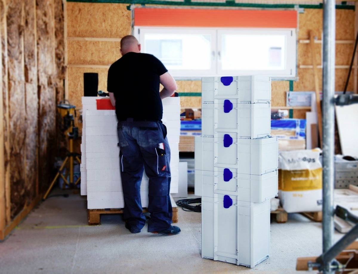 Osoba stojící v dílně vedle navrstvených strojních součástí, obklopená stavebními materiály a nástroji, s oknem v pozadí.