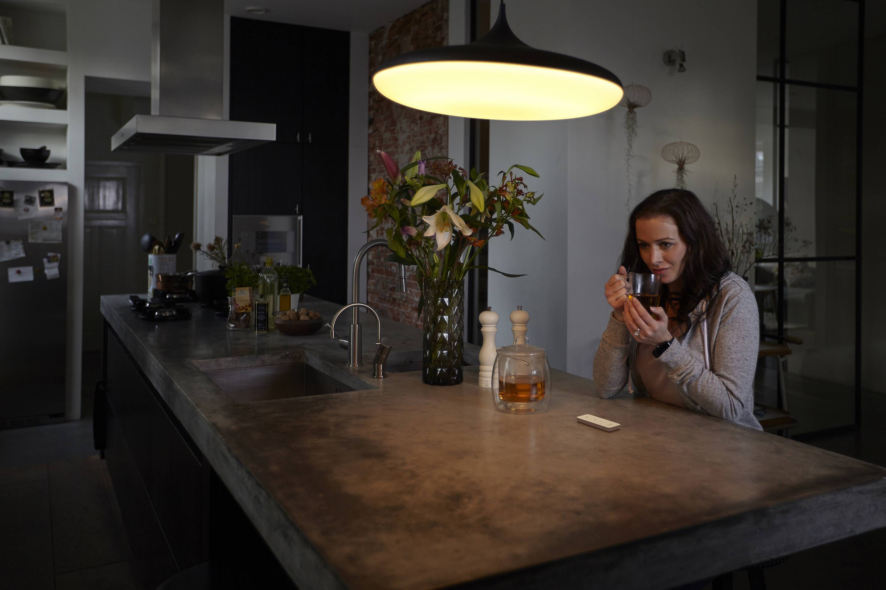 Žena sedí v moderní kuchyni u pultu, drží šálek a zamyšleně se dívá. Stůl s osvětlením, květinami a konvicí na čaj.