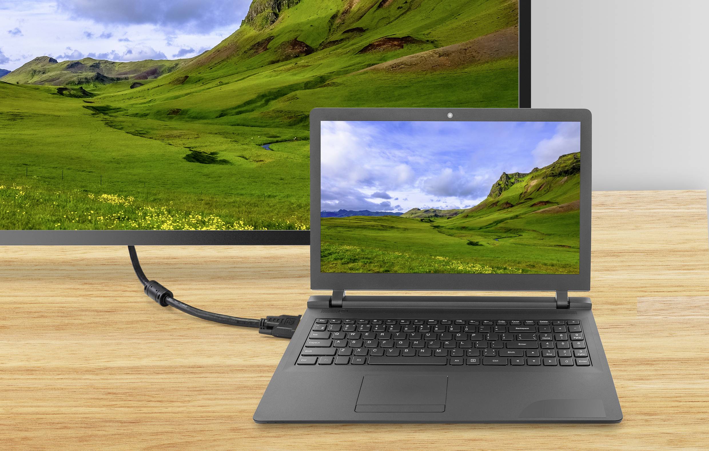 Notebook připojený pomocí HDMI kabelu k většímu monitoru, přičemž oba zobrazují obrázek zelených kopců a mraků.