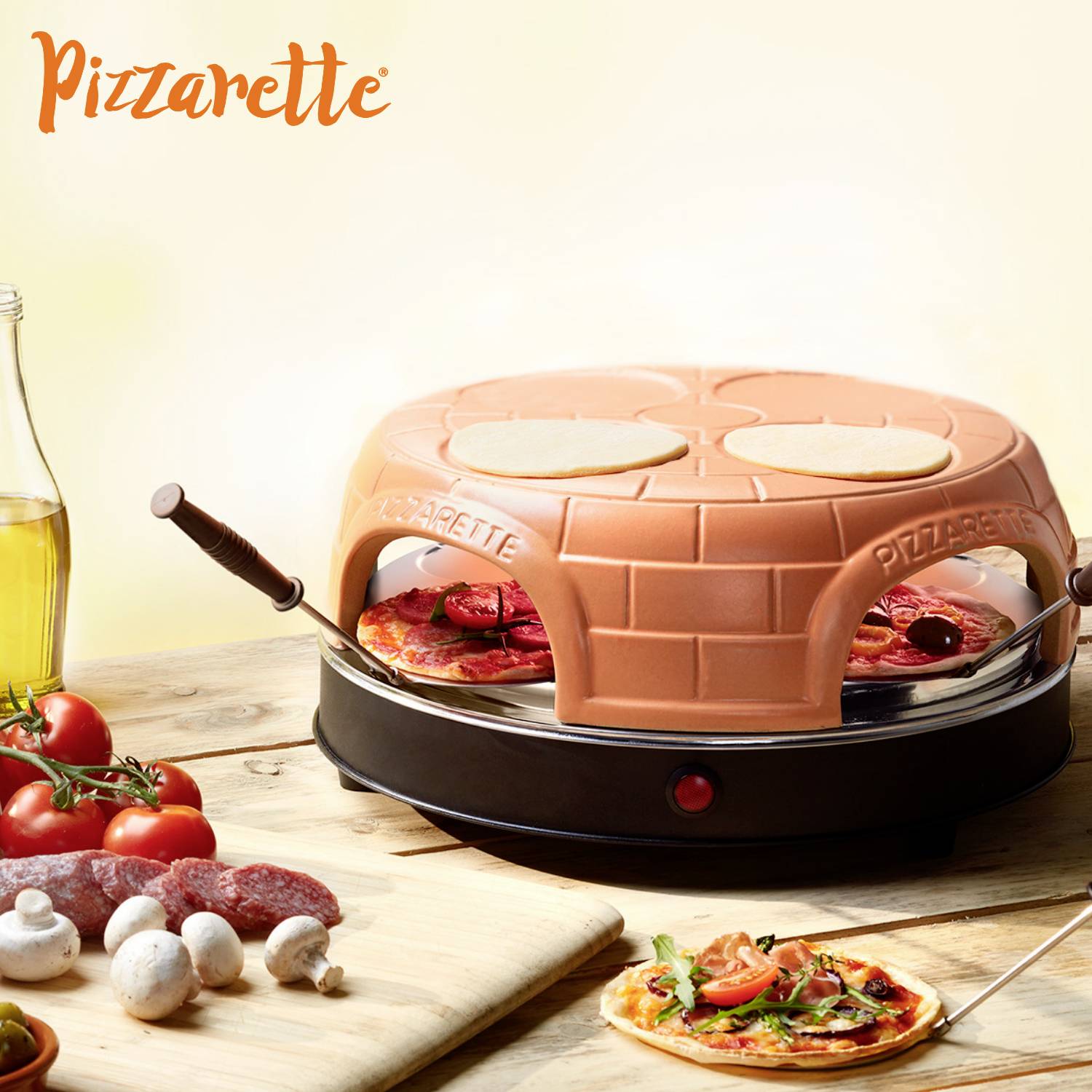 Miniaturní pizzová pec s terakotovou klenbou, obklopená ingrediencemi jako jsou rajčata, uzenina a talíř s hotovou pizzou.