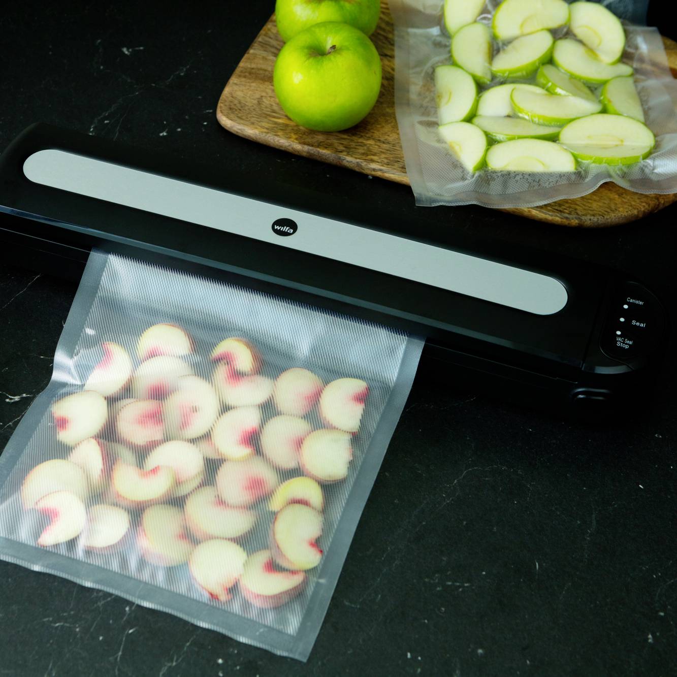 Vakuovací přístroj uzavírá sáček s nakrájeným ovocem. Vedle leží dřevěné prkénko s zelenými jablky a dalším nakrájeným ovocem.