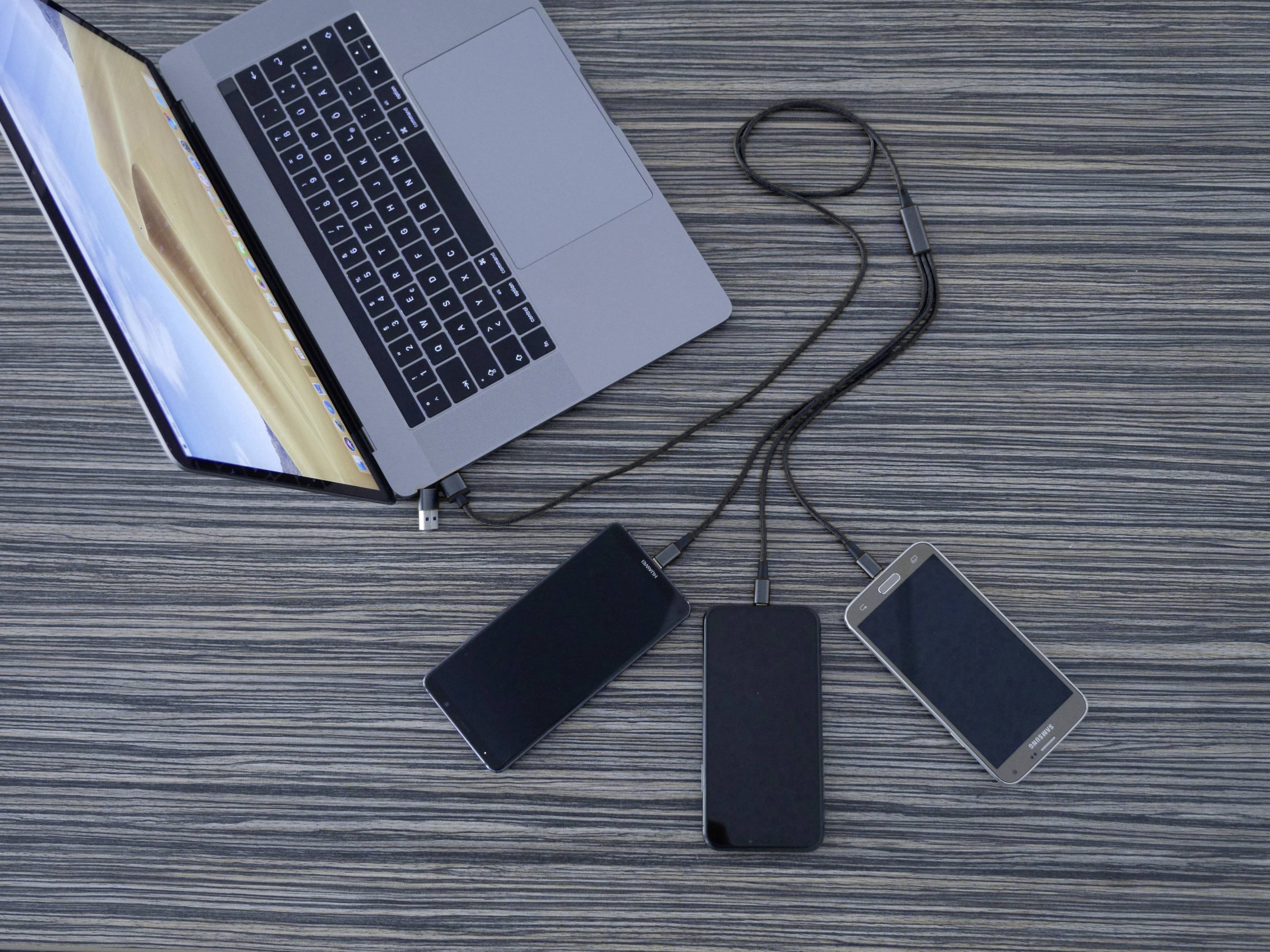 Laptop je připojen kabely k dvěma powerbankám a smartphonu a leží na stole s dřevěnou texturou.