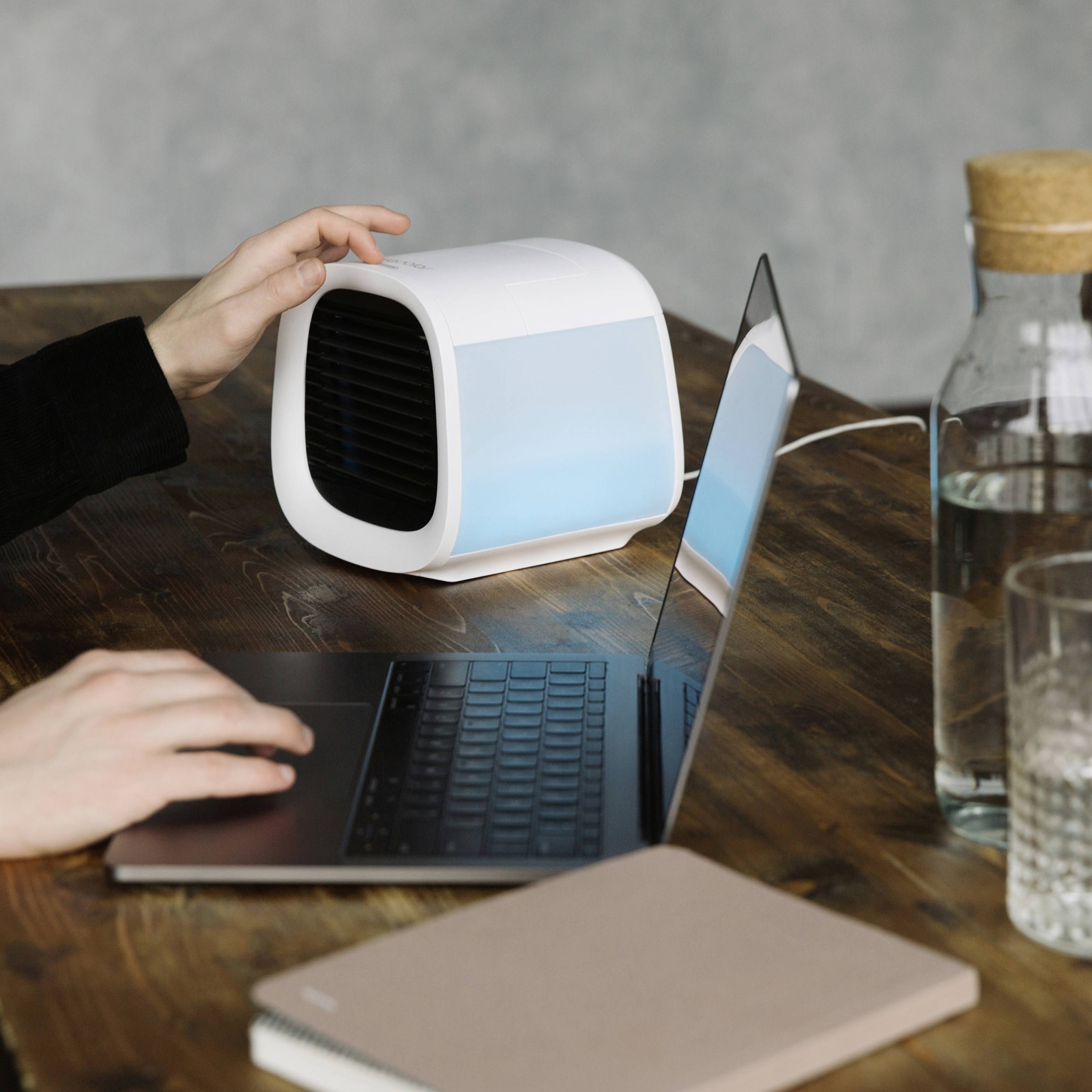 Člověk pracuje na laptopu u stolu. Vedle něj stojí malý, kubický klimatizační ventilátor. Poznámkový blok leží vedle laptopu.