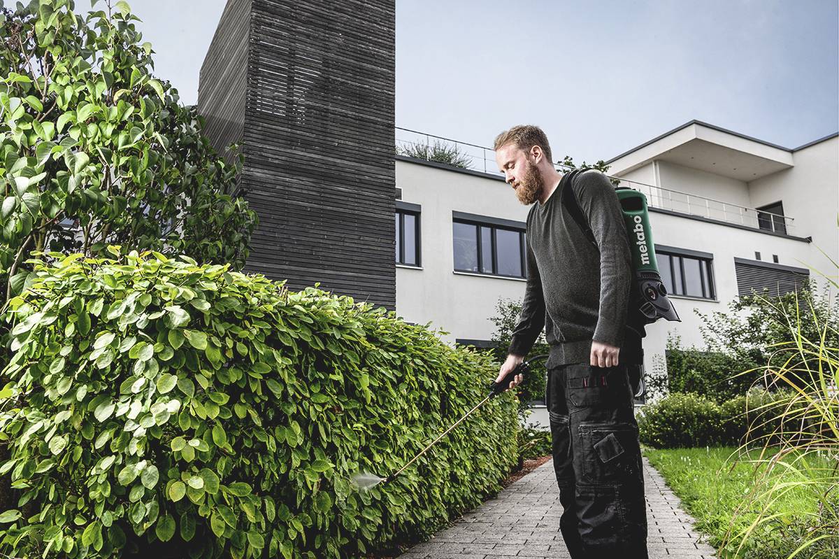 Muž s pomocí zahradního náčiní udržuje živý plot před moderní budovou. Obloha je jasná a prostředí působí upraveně.