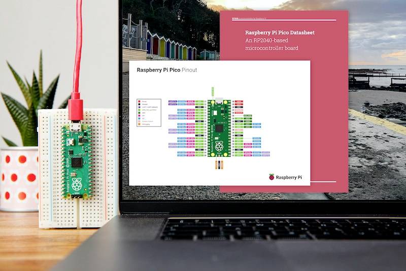 Laptop s otevřenou webovou stránkou s pinout diagramem Raspberry Pi Pico na obrazovce. Vedle něj namontovaný Raspberry Pi Pico na desce plošných spojů.