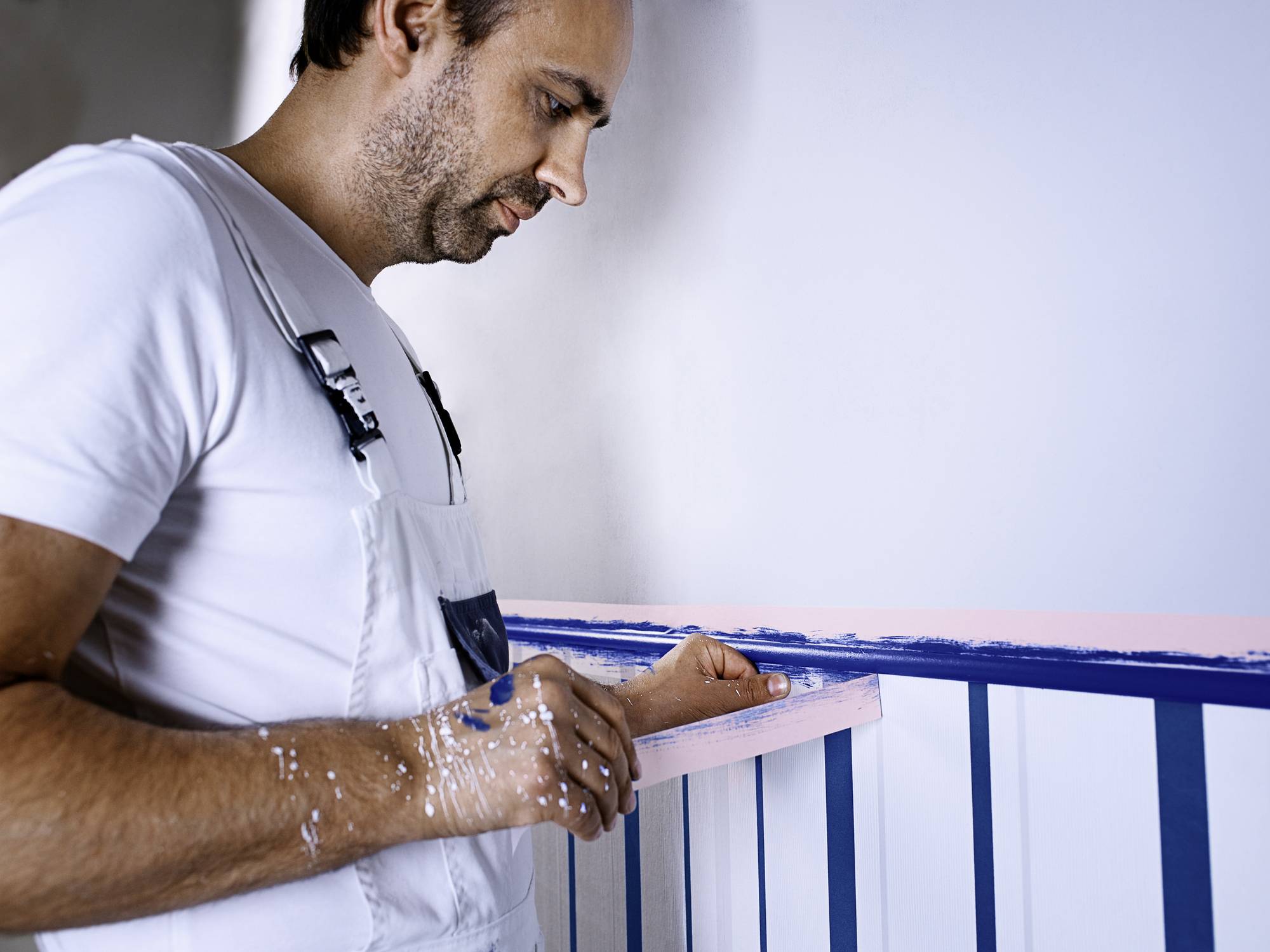 Muž v bílém overalu maluje zábradlí na modro. Soustředěně pracuje, s modrými skvrnami od barvy na rukou.